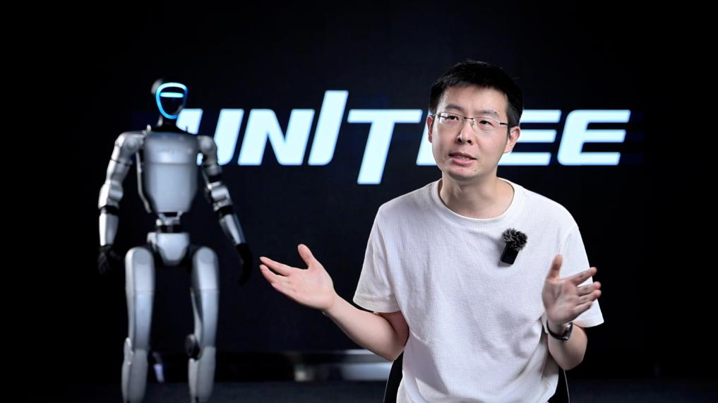 Wang Xingxing, founder of Unitree, speaking on stage beside a humanoid robot