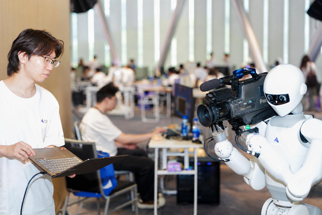 A developer with a laptop beside a humanoid robot carrying a broadcast camera at the Shenzhen hackathon final